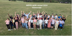 A diverse group of PhD students standing on a grassy field, smiling and waving at the camera. Text reads "IMPRS-MOB retreat 2024 Hohenroda."
